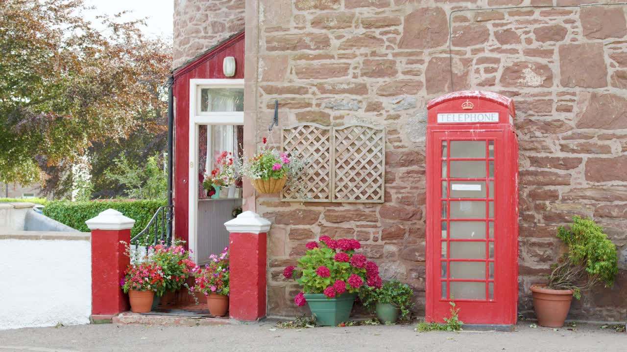 Classic red phone booth beside stone house with colorful potted flowers in soft daylight