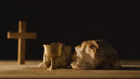 concepto religioso filmado con cruz de pan roto cáliz y vino en mesa de madera contra fondo negro