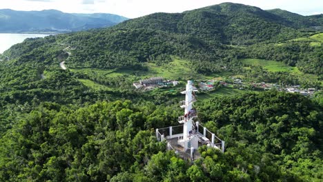 Cinematic-Aerial-View-of-White-Lighthouse-on-top-of-mountain-in-lush-tropical-jungle-with-small-town-in-background