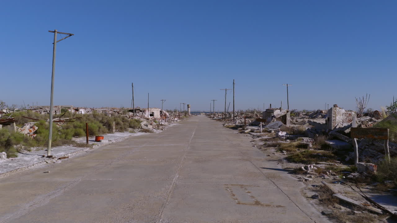 Drone a bassa quota avanza lungo una strada a Epecuén, affiancato da rovine di edifici con acqua di fronte
