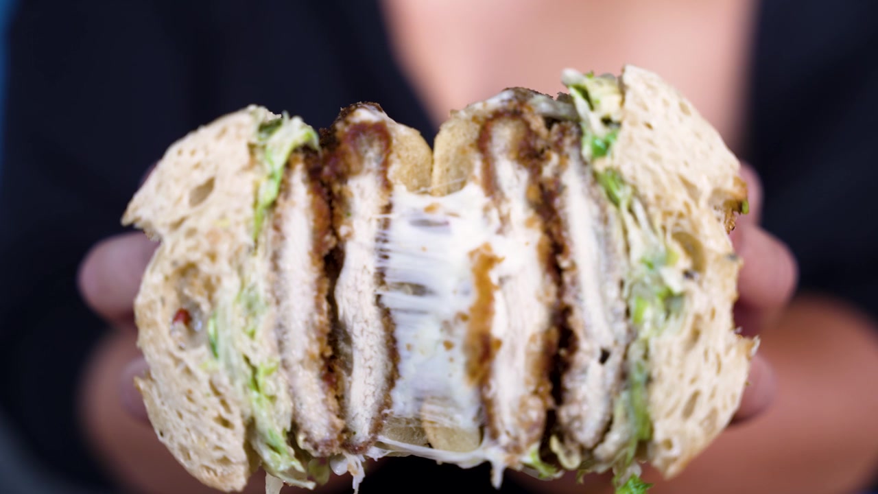 Primo piano in 4K di un panino con cotoletta di pollo, avocado e formaggio filante che viene aperto da mani con una manicure perfetta per rivelare il ripieno