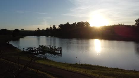 Movimiento-Hacia-Adelante-Con-Drone-Hacia-Un-Gran-Lago-Con-Puesta-De-Sol-En-El-Fondo,-Cielo-Azul-Y-Luz-Brillante-Con-Reflejos