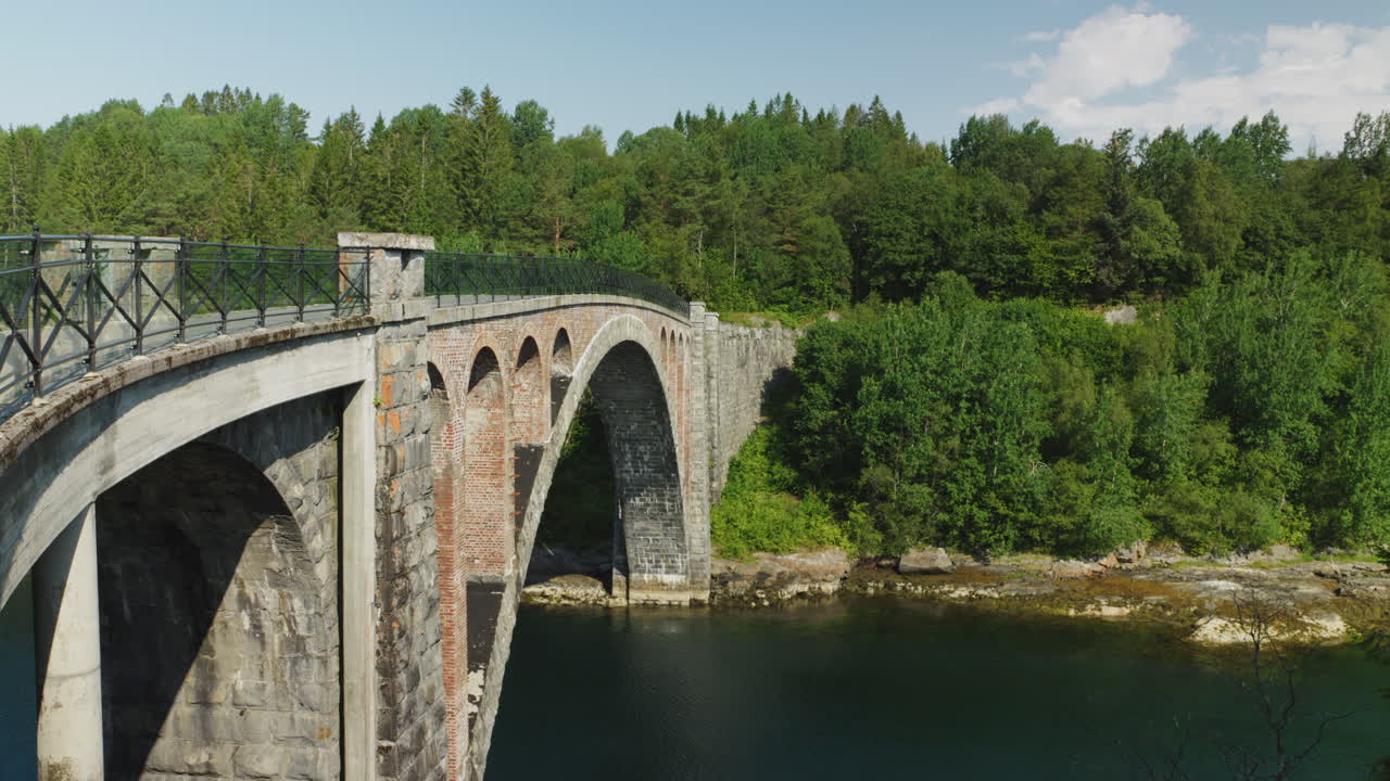 Wide shot of an old bridge in Norway, surrounded by nature and trees and the water flows calmly under it on a bright sunny day
