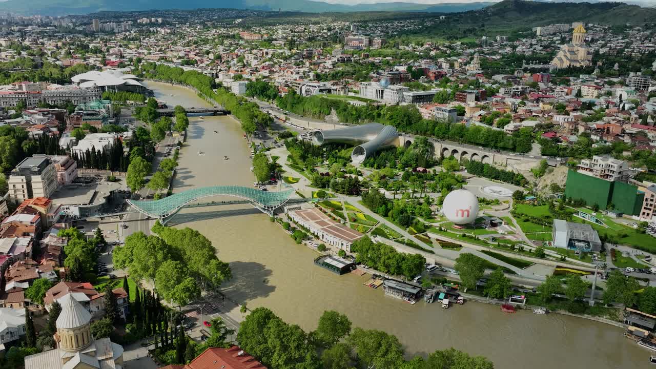 A modern aerial view featuring the Peace Bridge, Rike Park, and the flowing Kura River surrounded by the vibrant urban center of Tbilisi