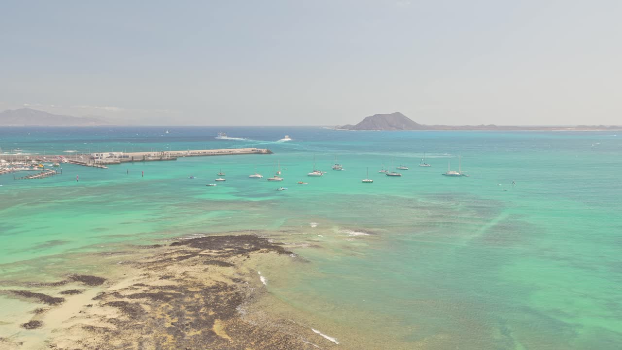 Aerial panorama showing sailboats navigating turquoise waters beside Playa del Medio, Fuerteventura, with Isla de Lobos silhouetted against horizon