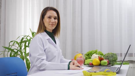 mujer dietista positiva trabajando en una mesa llena de verduras y frutas sonriendo a la cámara.