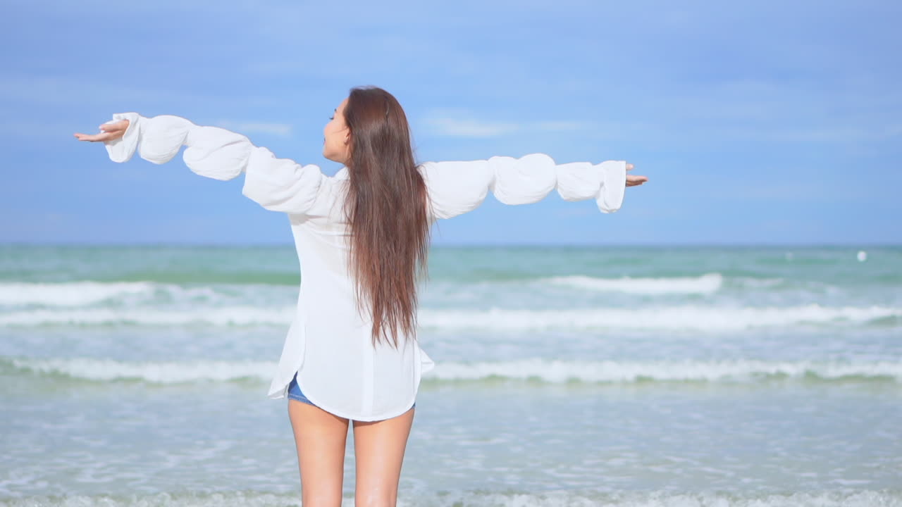 카메라에 등을 대고 있는 젊은 여성이 팔을 공중으로 들어 올려 들어오는 서핑을 향해 걸어갑니다.