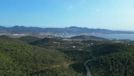 road winding through a vibrant green forest in ibiza, spain, leading down towards the beautiful san antonio bay. great aerial view flight static tripod hovering drone