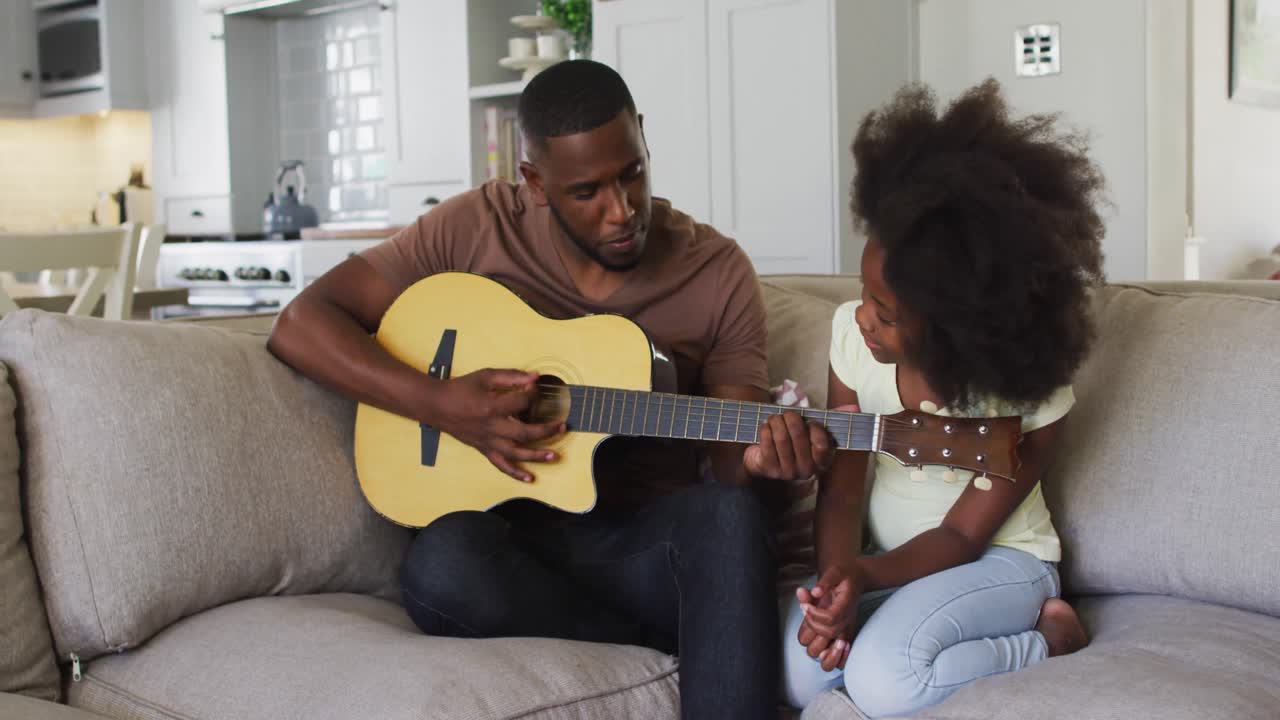 Padre afroamericano che suona la chitarra seduto sul divano con sua figlia.