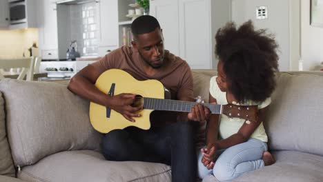 African-american-father-playing-guitar-sitting-on-couch-with-his-daughter