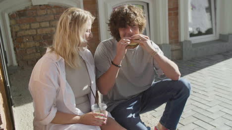 young couple sharing a tasty hambuger, sitting on the sidewalk in the city