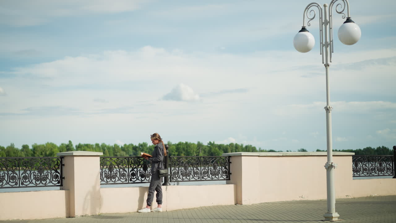mujer con ropa gris y bolso negro se encuentra cerca de la valla en la orilla del río, leyendo un libro, los árboles son visibles en la distancia, y un poste de luz se encuentra cerca