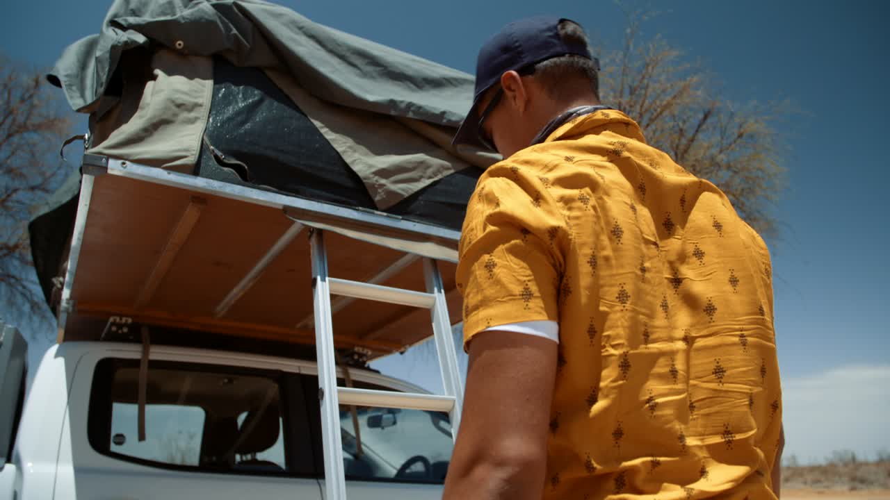 Hand held shot of a tourist climbing up a rooftop ladder and starts to roll up his canvas tent door.