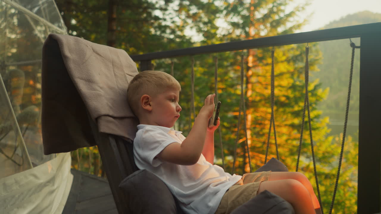 Boy plays game on mobile phone sitting in armchair on balcony with incredible view of nature beauty. Luxurious glamping in country house slow motion