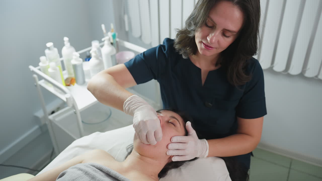 Specialist wearing surgical gloves dips fingernail inside guest mouth and cradles jawline during cheek massage as guest closes eyes and relaxes on massage chair in clinical setting