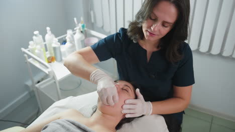 specialist wearing surgical gloves dips fingernail inside guest mouth and cradles jawline during cheek massage as guest closes eyes and relaxes on massage chair in clinical setting