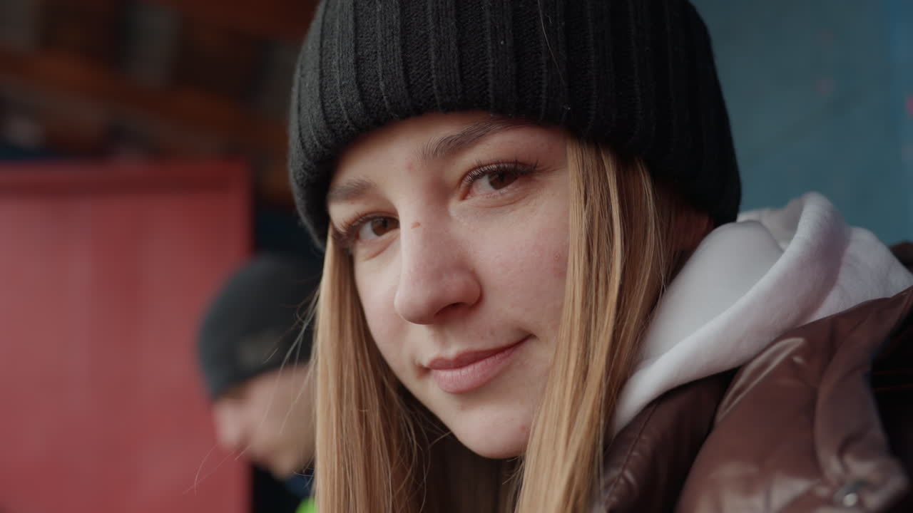 흰색 옷을 입은 여성이 녹색 재킷, 비니, 갈색 패딩을 입은 남성 동반자 옆을 내려다보고 있어요. 멋진 도시 배경에 부드러운 표정과 사려 깊은 분위기가 가까이에서 포착된 친밀하고 솔직한 인물 사진입니다.