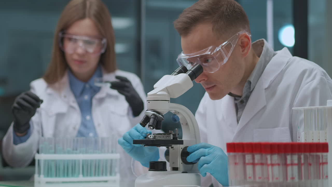 микробиологические исследования двух экспертов в медицинской лаборатории мужчина и женщина исследуют образцы с помощью пробирки и микроскопического аппарата