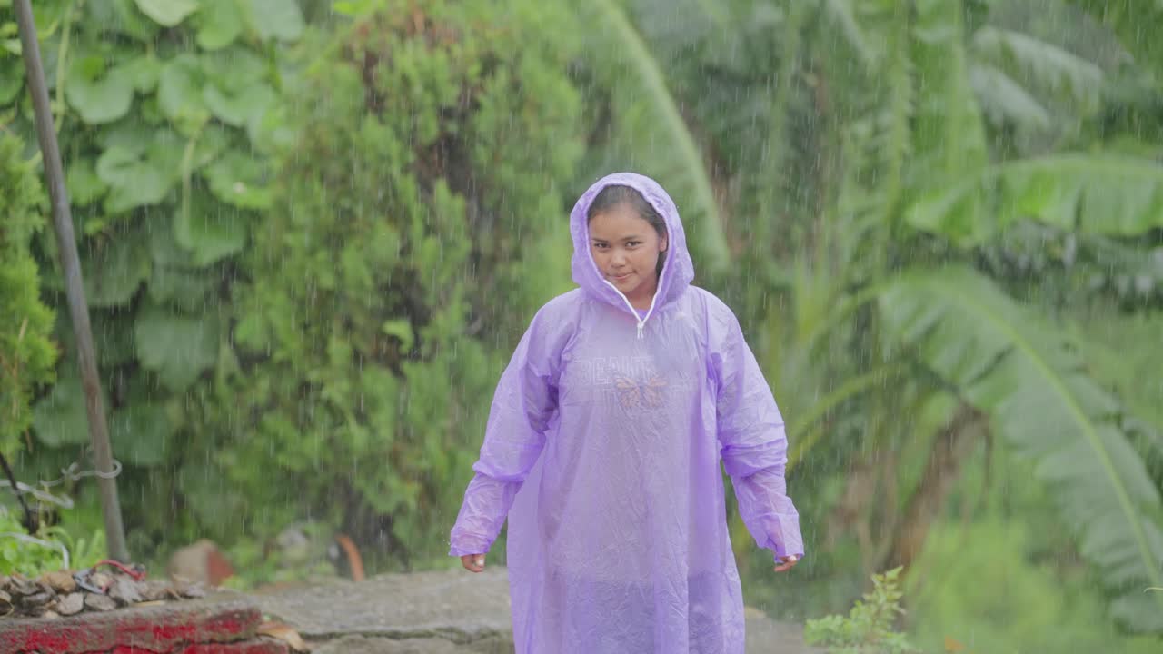 Happy Indian girl in raincoat playing in the monsoon rain, cheerful expression in village environment, 4k video