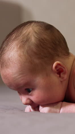 infant lying on the bed on his belly. sweet boy chewing his tiny fists. little kid lying on the bed in a light room close up. vertical video