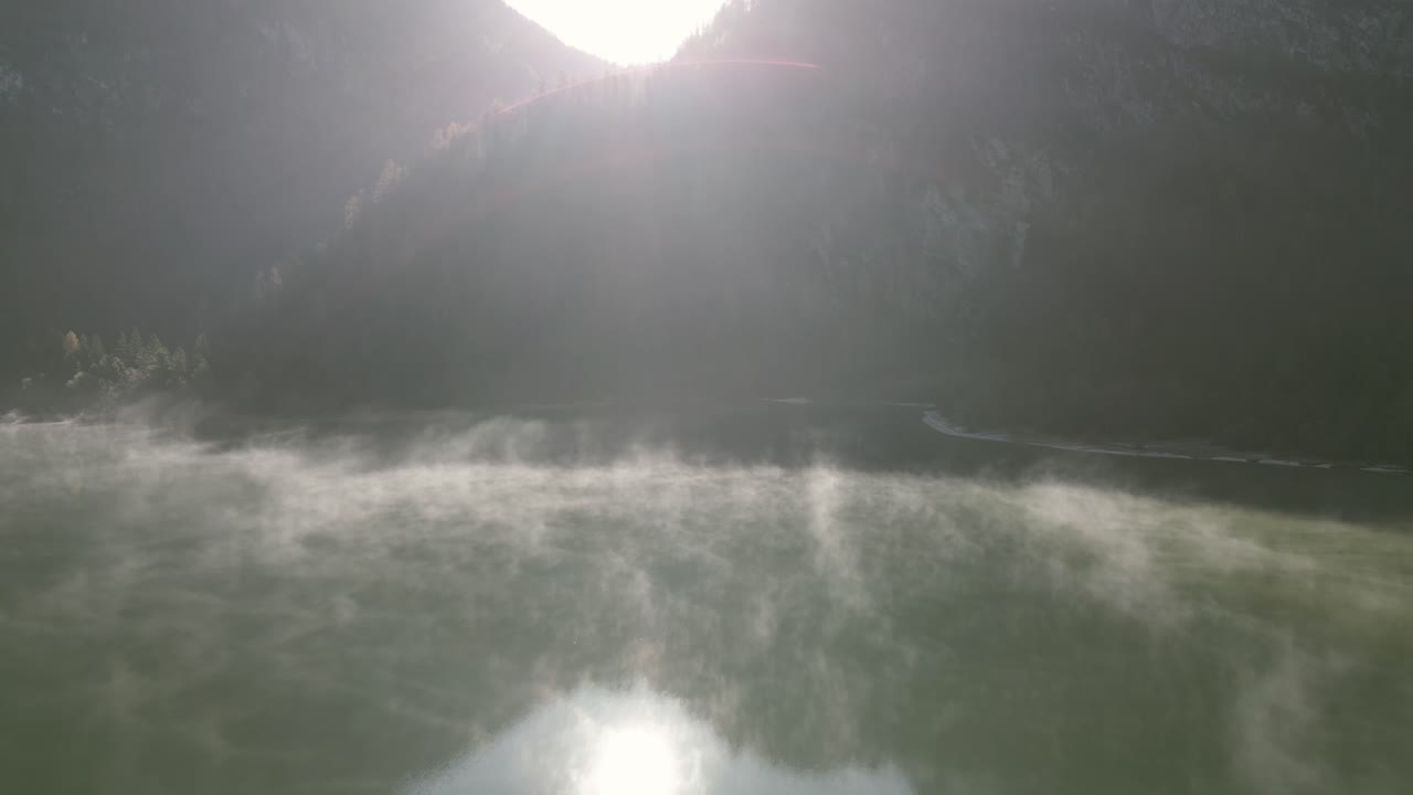 Drone view over Predil Lake, Slovenia: turquoise waters reflect the surrounding mountains, with lush green forests lining the shore. The serene lake is nestled in a valley