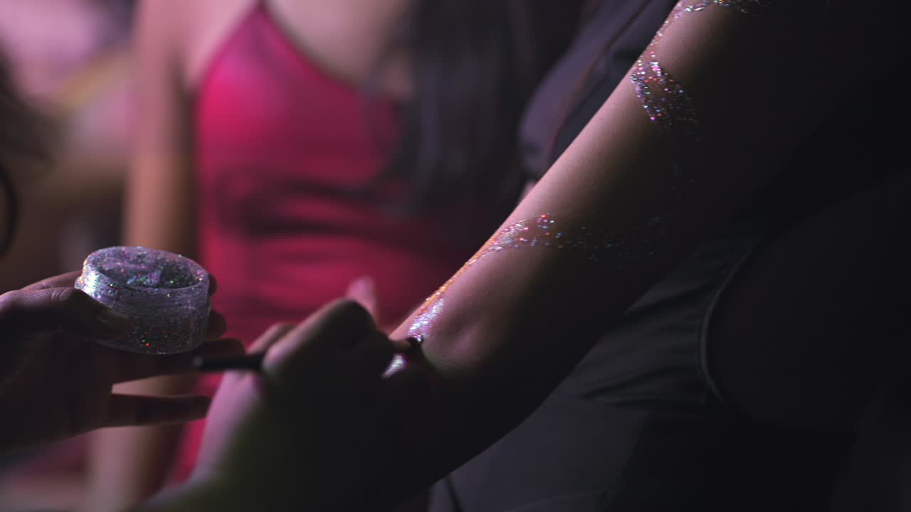 Shallow depth of field video showing a girl painting another's arm with glitter. Other girls wait in line at a night party in Argentina, in 4K