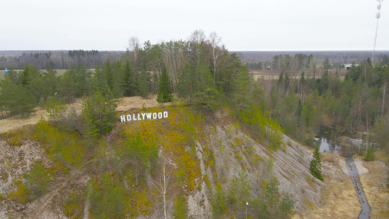 Aerial drone view orbiting and spinning around a large white Hollywood sign during a cold or chilly cloudy autumn or spring day in Estonia. The sign is high up on a hill with trees growing on gravel.