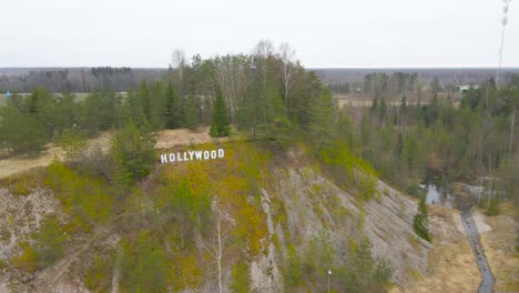 aerial drone view orbiting and spinning around a large white hollywood sign during a cold or chilly cloudy autumn or spring day in estonia. the sign is high up on a hill with trees growing on gravel.