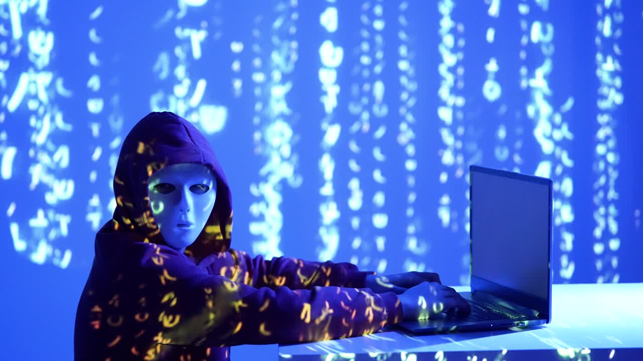 A hooded hacker in a white mask stares ahead as police-style red and blue lights flash across binary code. The scene evokes cybercrime, tension, and the anonymity of the digital underworld