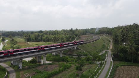 drone shot, indonesian train passing through a double track railway bridge winding on the left and right with rice fields, forests and villages.