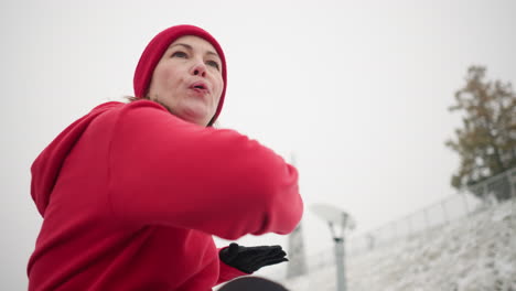 close-up of lady exercising outdoors, raising hands for balance, turning left and right in winter setting with snowy hill, iron railing, blurred light pole, and vibrant red hoodie for warmth