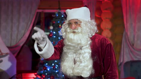 portrait of a real santa with white beard. senior man in santa clause costume wearing glasses standing in christmas decorated room and talking. christmas spirit, traditions concept