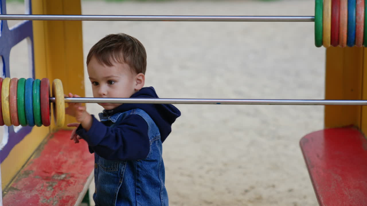Little toddler wearing jeans jacket plays at the playground. Cute child moving rings on the big abacus and runs away.