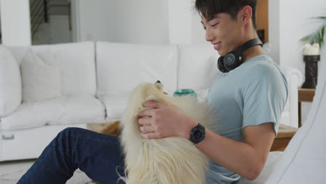 행복한 아시아 남성 십대가 거실에서 개를 키우고 헤드폰을 착용하고 있습니다.