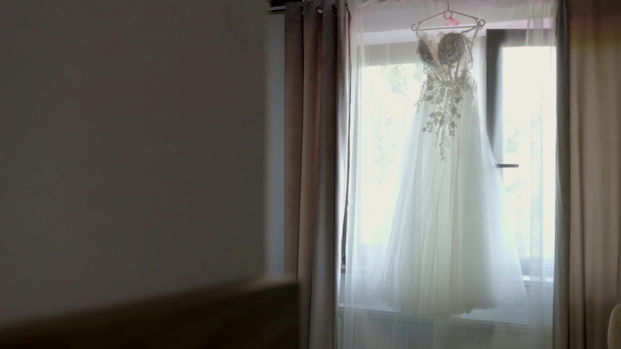 A beautiful wedding dress hanging on a hanger by the window, peeking out from behind the corner of the wall.