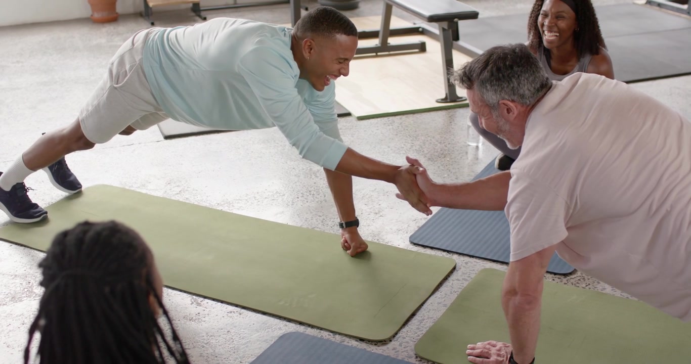 Lezione di fitness multietnica che fa plank, un uomo afroamericano con maglietta blu tocca il partner sui tappetini per l'equilibrio.
