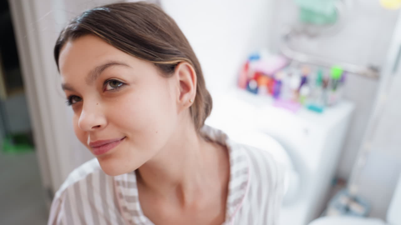 Donna caucasica con pelle fresca che sorride al suo riflesso nello specchio del bagno, donna gioiosa che mostra un incarnato luminoso dopo la detersione mentre sorride in un bagno ben illuminato