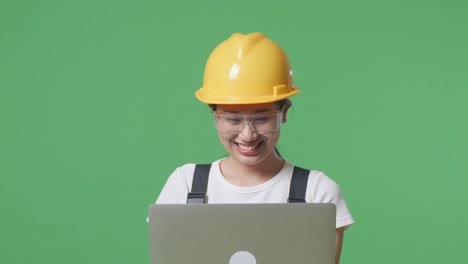 close up de una trabajadora asiática que usa gafas de protección y casco de seguridad usando una computadora portátil y sonriendo mientras está de pie en el estudio de fondo de la pantalla verde