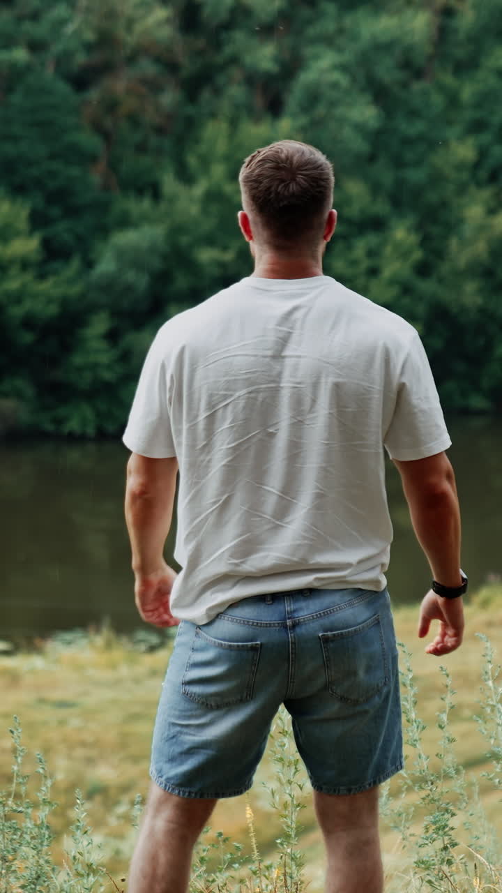 Rear view of a sportive muscular man in white t-shirt and jeans shorts. Happy person in the nature surrounding enjoying life. Vertical video
