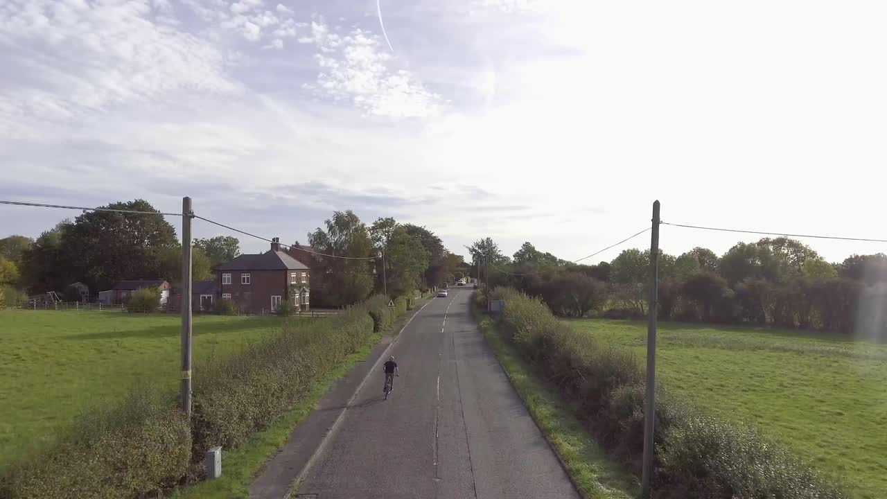 A Biker rides into a village in the UK.  Drone shot where the camera gradually gets higher and reveals the rural landscape.