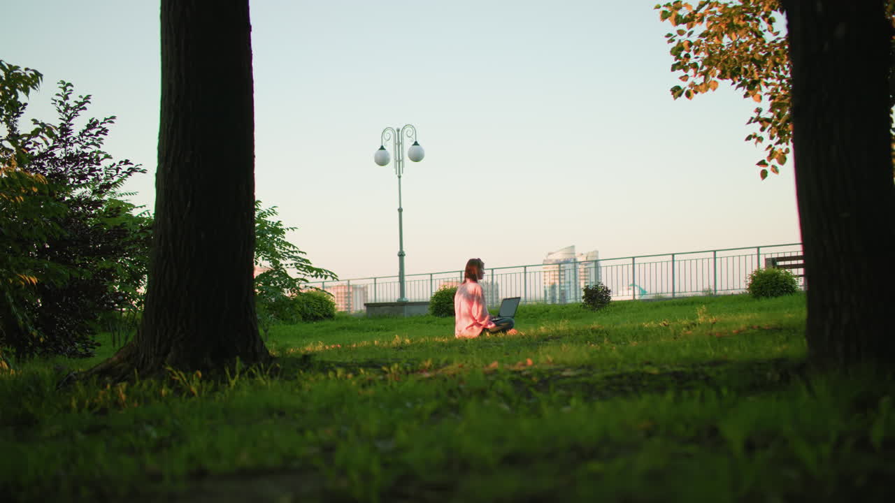 잔디에 앉아 노트북으로 일하는 여성, 배경에는 조명 기둥, 철로, 도시 건물, 도시 요소와 자연을 결합하는 평화로운 설정