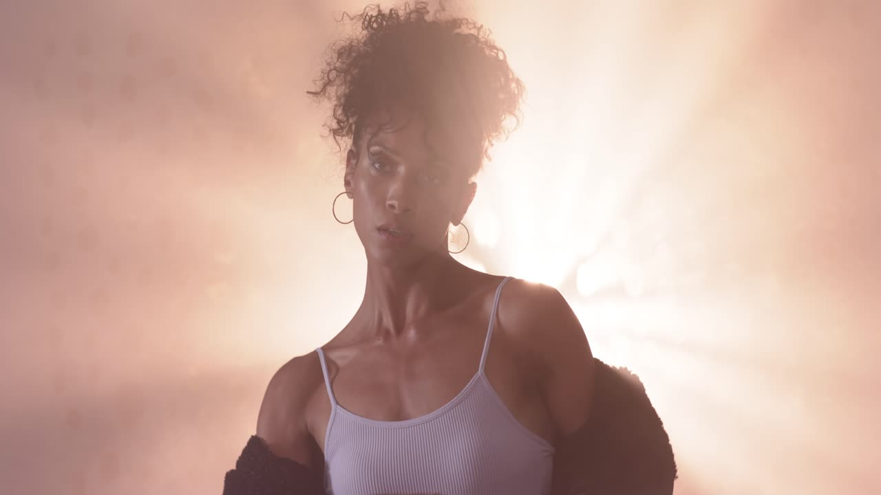 Uma mulher afro-americana, ao sinal, tira a jaqueta dos ombros e ajusta a blusa para uma sessão de fotos em estúdio com névoa.