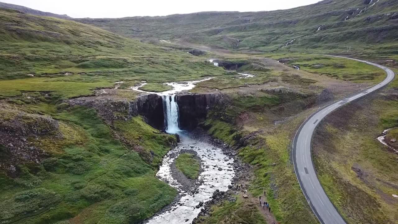 A breathtaking aerial view of a cascading waterfall nestled in green hills, with a curving road weaving through the picturesque landscape of Grabrok, Iceland. A serene natural escape