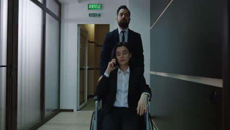 Wheelchair Manager Phoning in Office Corridor. Medium shot of disabled manager phoning while colleague pushing her wheelchair in office corridor