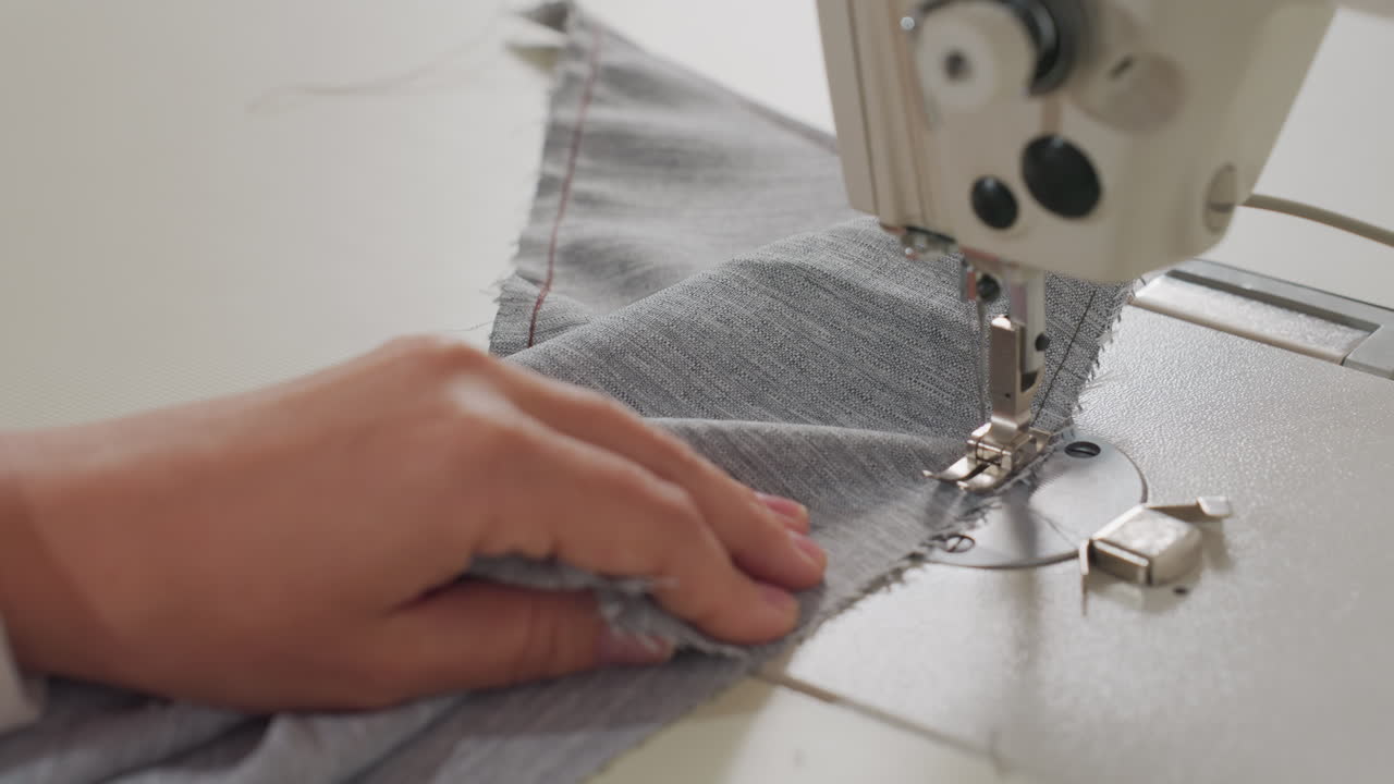 Female hand gently guides fabric under needle and thread machine during garment stitching process, showcasing careful technique and textile control in soft daylight on white sewing surface