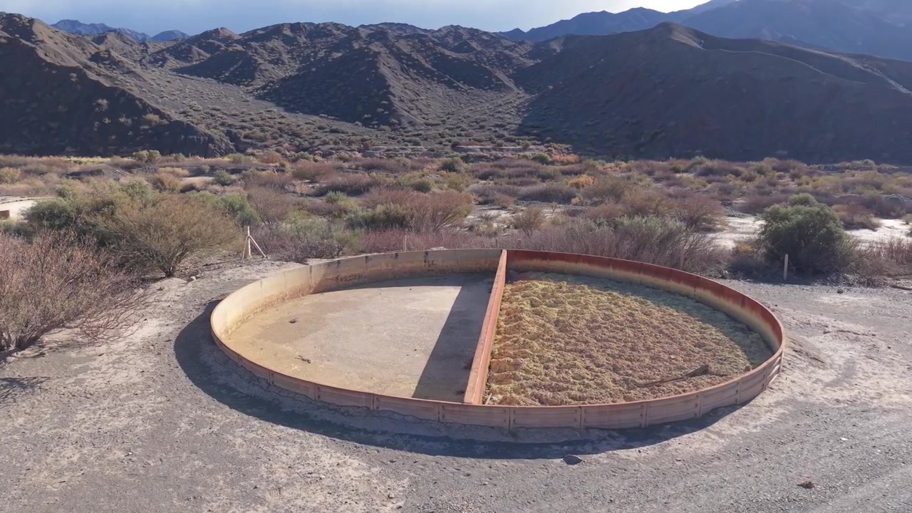 Drone video captures the abandoned circular tank of Planta de Sulfato de Aluminio in San Juan, Argentina, set against a dramatic arid mountain backdrop, early morning natural light.