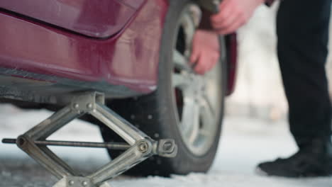 close up of car jack positioned under vehicle on snowy ground, person approaching to loosen rim bolt, clear focus on jack, tire, and effort in outdoor winter setting for car maintenance