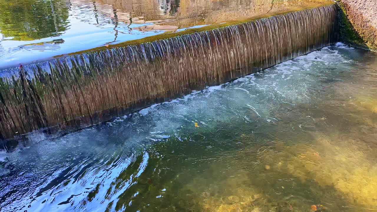 L'acqua scorre su una serena diga sotto la luce intensa del sole, creando increspature e riflessi in un ambiente naturale pacifico circondato dalla vegetazione.