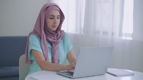 una joven musulmana hermosa está trabajando en una computadora portátil en su lugar de trabajo. una joven musulmana sentada frente a la pantalla de una computadora portátil mira hacia arriba y mira a la cámara web