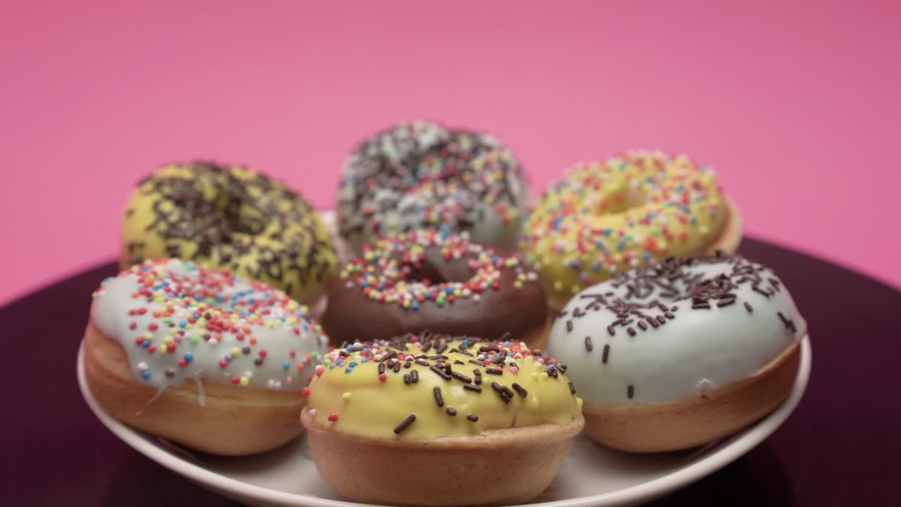 A bunch of American donuts with black, yellow and blue chocolate and choco and multicolored crumb toppings rotates on a pink and black background in a curcle